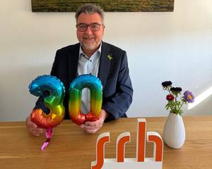 Ein Mann sitzt mit zwei Luftballons in Form der Zahl 30 und hält sie. Hinter ihm hängt ein Landschaftsbild, daneben steht eine Vase mit Blumen.