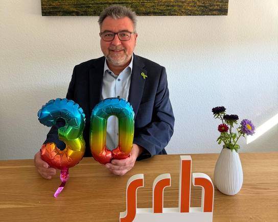 Ein Mann sitzt mit zwei Luftballons in Form der Zahl 30 und hält sie. Hinter ihm hängt ein Landschaftsbild, daneben steht eine Vase mit Blumen.