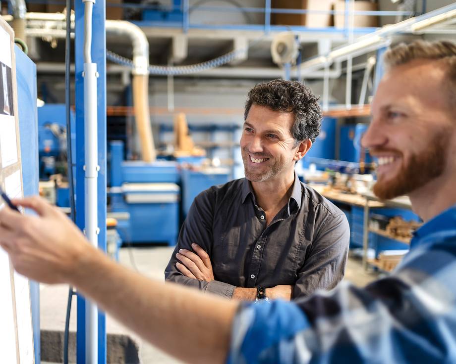Zwei Männer stehen in einer industriellen Umgebung und schauen auf ein Whiteboard.